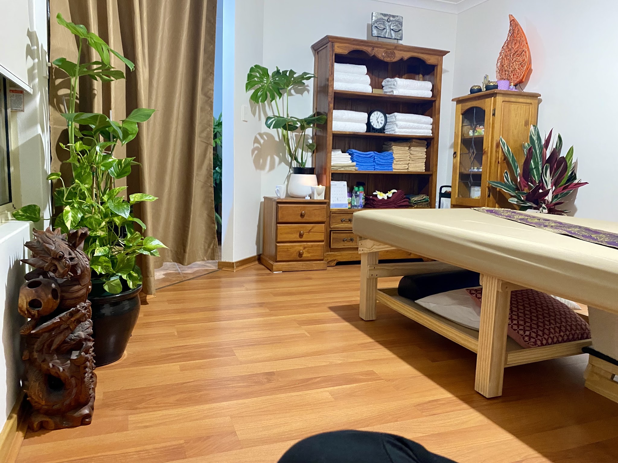 Wide view of the treatment room with timber cabinets, towel shelves, indoor plants and the massage table